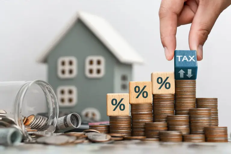 Una mano posiziona un cubo con la scritta «TAX» in cima a una pila di monete vicino a un modellino di casa, per illustrare il concetto di imposta sugli immobili.