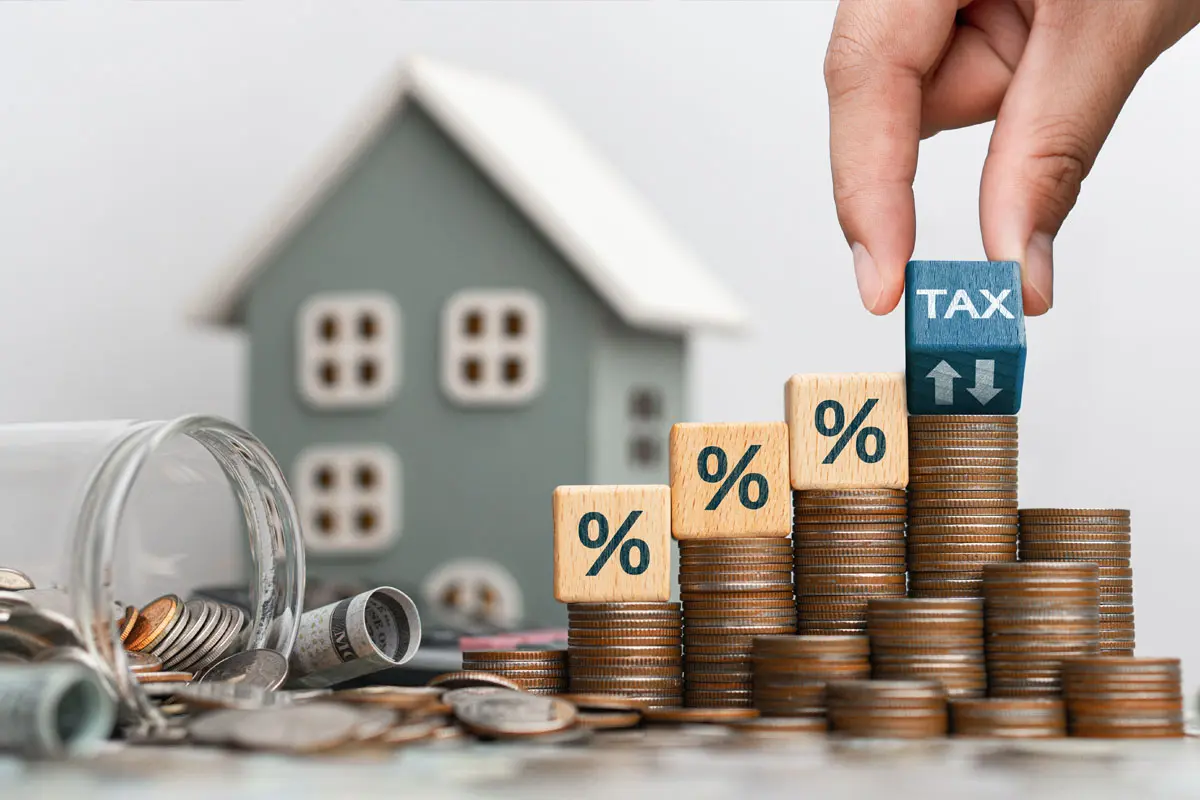 Una mano posiziona un cubo con la scritta «TAX» in cima a una pila di monete vicino a un modellino di casa, per illustrare il concetto di imposta sugli immobili.